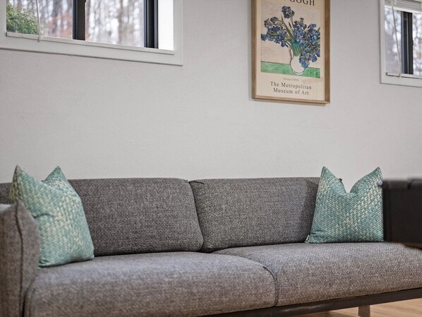 A grey sofa with two pillows, a framed artwork above it, and a window to the left.