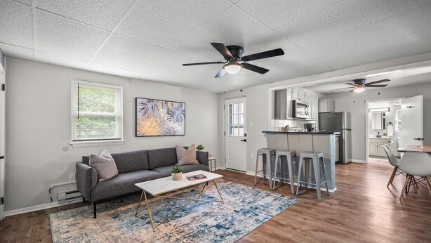 A spacious living area with a gray sofa, coffee table, and a blue patterned rug. A ceiling fan and kitchen are visible in the background.
