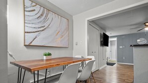 A modern dining area with a wooden table and chairs, a large abstract painting on the wall, and a kitchen area in the background.