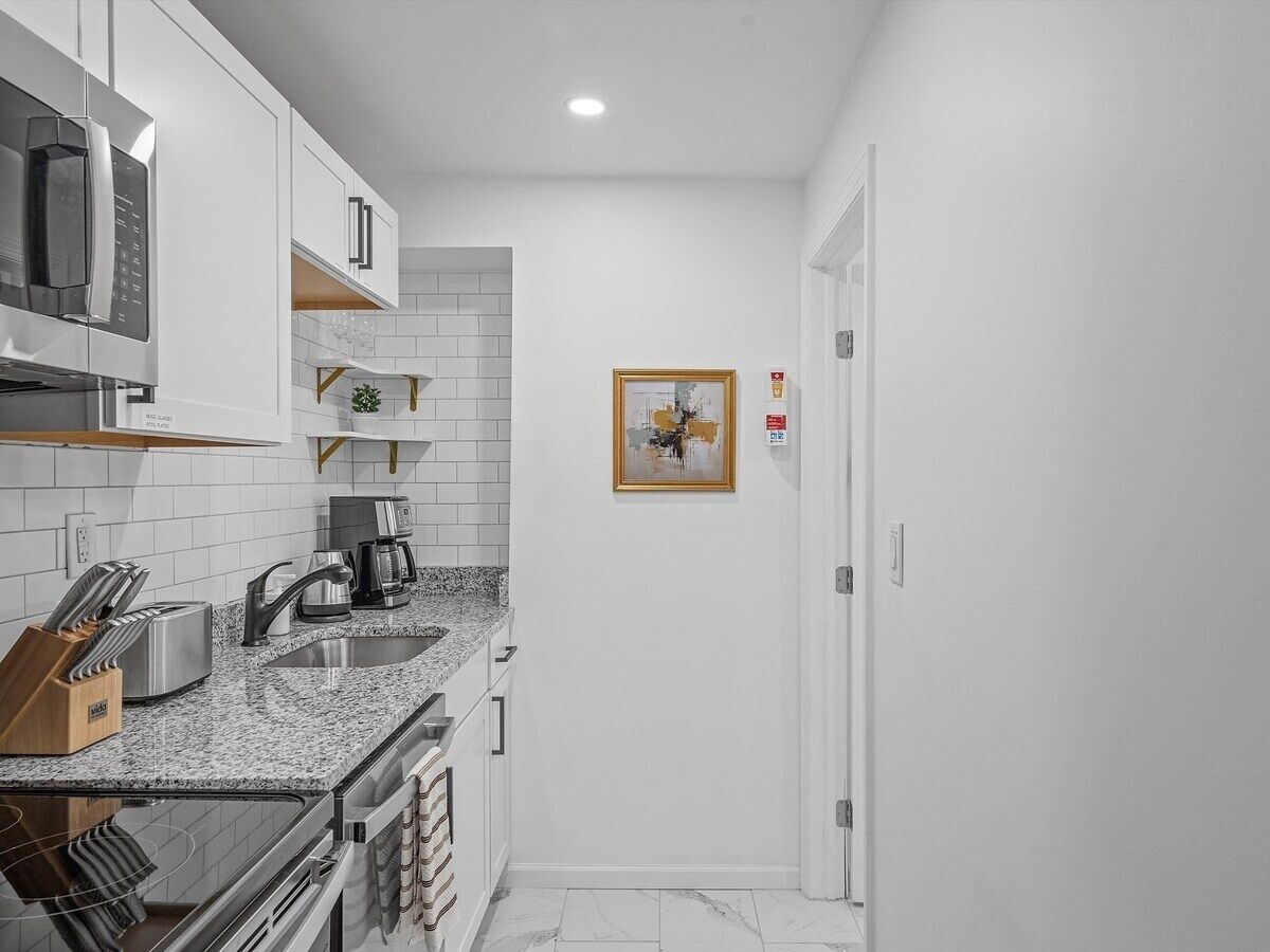 A compact kitchen with white cabinets, a granite countertop, a microwave, a coffee maker, and a framed picture on the wall.