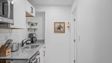 A compact kitchen with white cabinets, a granite countertop, a microwave, a coffee maker, and a framed picture on the wall.