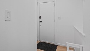 Un couloir étroit avec une porte blanche, un paillasson noir et une étagère blanche.