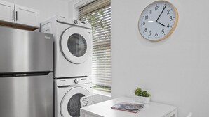 Cocina compacta con refrigerador de acero inoxidable, lavadora y secadora apiladas, una mesa blanca con una planta y un reloj de pared.