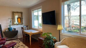 A living room with a dining table, chairs, a sofa, a television, and a large window with a view of the outdoors.