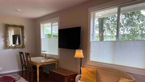 A room with a bed, a dining table with chairs, a television, a lamp, and a wicker sofa with cushions.