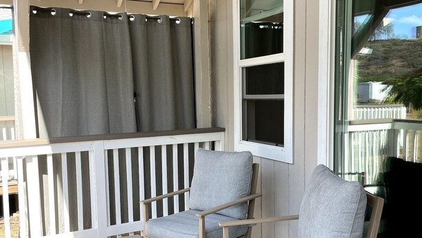 Un porche abrité avec deux fauteuils gris à revêtement en tissu et une petite table, des balustrades en bois et une fenêtre avec un rideau.