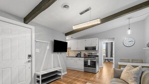Una cocina moderna con armarios blancos, horno empotrado y microondas. Hay una zona de comedor con una mesa y sillas, y una sala de estar con sofá y televisor.