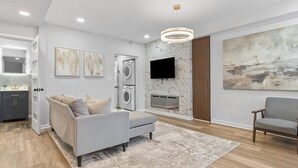 Een moderne woonkamer met een grijze bank, een salontafel, een flatscreen-tv aan de muur, een groot abstract schilderij en een ingebouwde wasmachine.