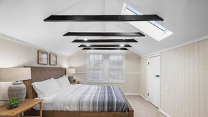 Un dormitorio con techo inclinado, tragaluces, estructura de cama de madera, ropa de cama blanca y una mesita de noche con lámpara.