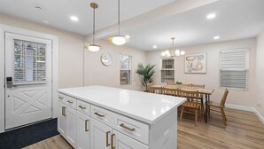 Una cocina moderna con armarios blancos, una isla central, iluminación colgante y un comedor con sillas y mesa de madera.