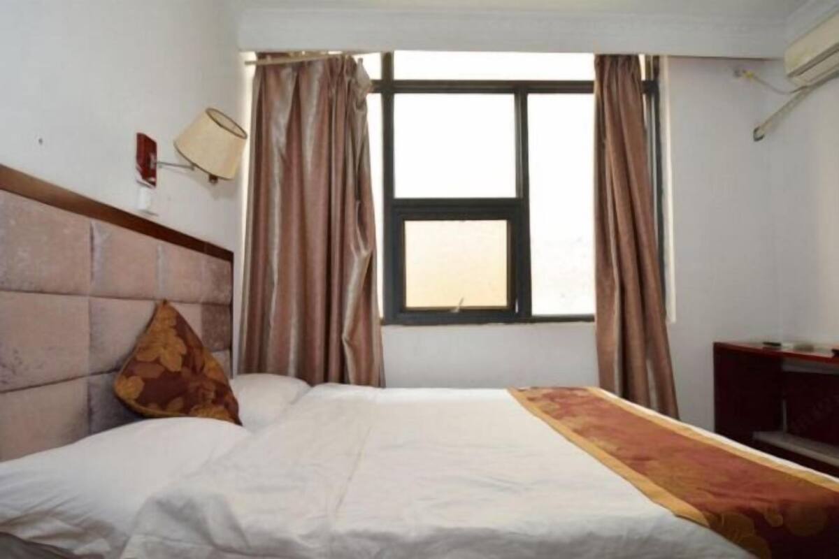 A hotel room with a bed, curtains, a window, and a wall-mounted lamp.