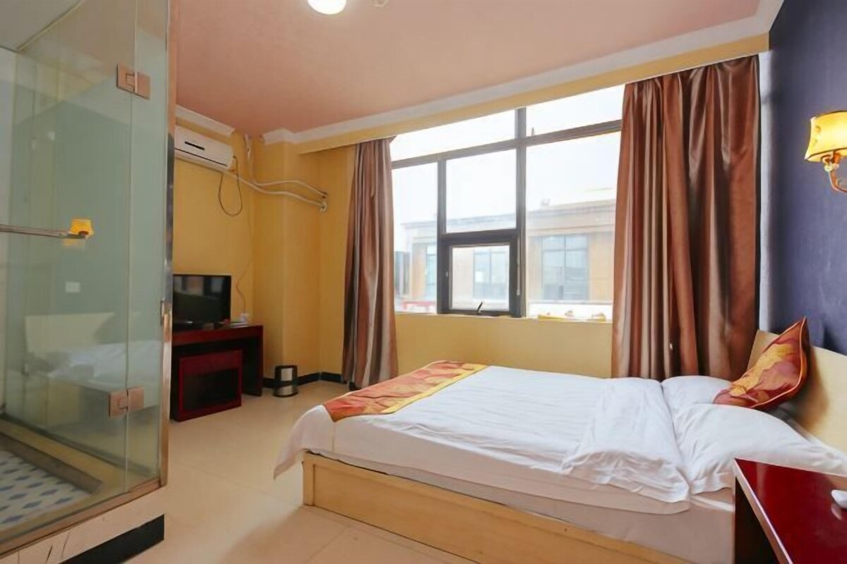 Habitación de hotel con cama, escritorio, televisión, cabina de ducha de vidrio y ventana con cortinas.