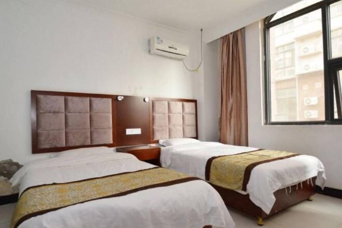 A hotel room with two beds, a window with curtains, an air conditioning unit, and a view of buildings outside.