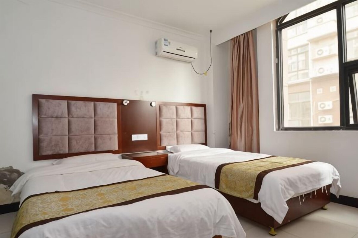 A hotel room with two beds, a window with curtains, and an air conditioning unit.