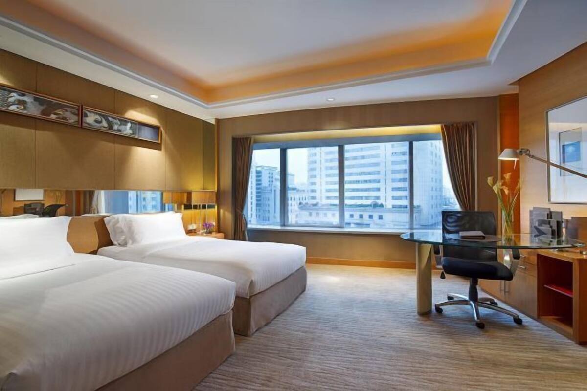 Une chambre d’hôtel avec deux lits, un bureau, une chaise, une grande fenêtre donnant sur la ville et un plafond doté d’un éclairage encastré.