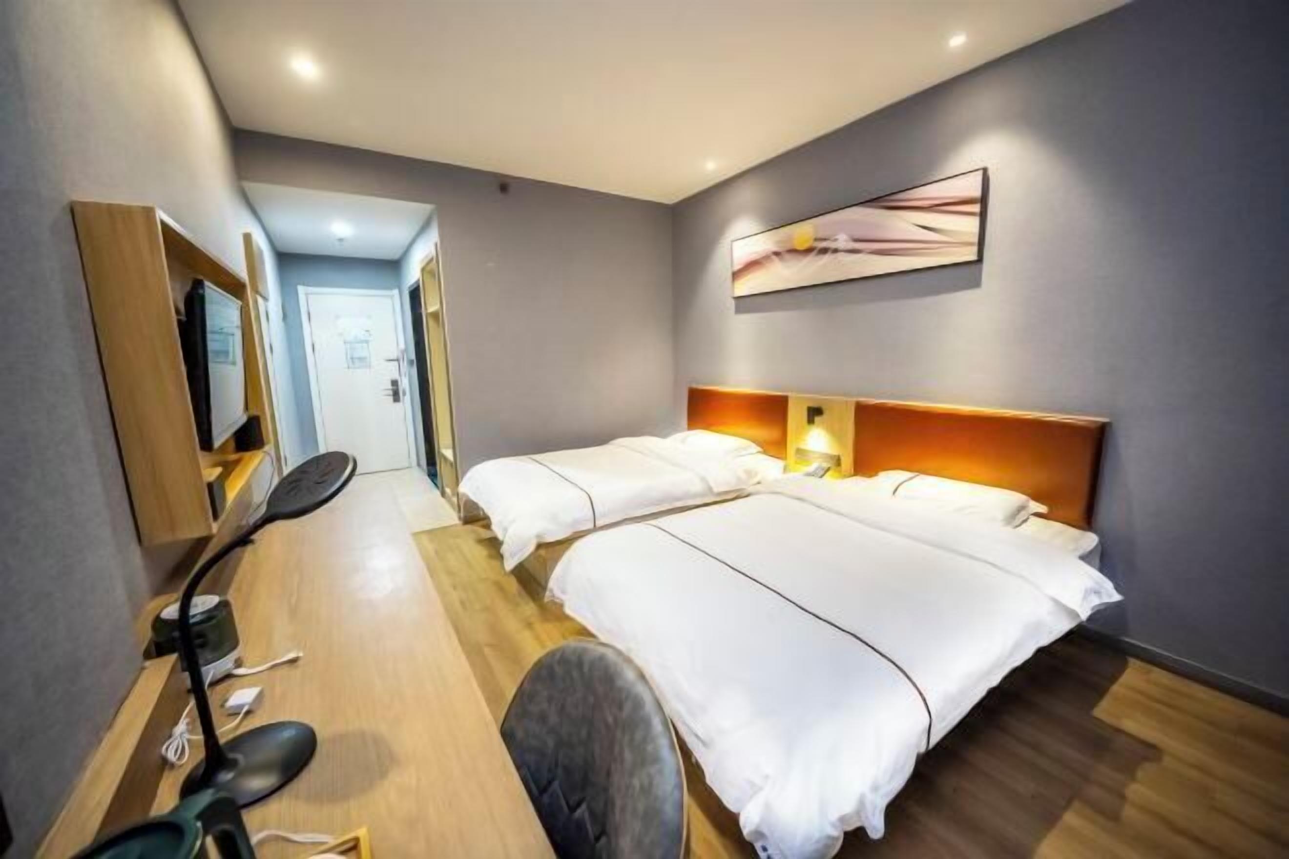 A hotel room with two beds, a desk with a chair, a desk lamp, and a wall-mounted TV.