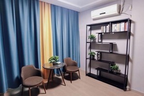 Una habitación con cortina azul y amarilla, una estantería negra con plantas y dos sillas alrededor de una pequeña mesa.