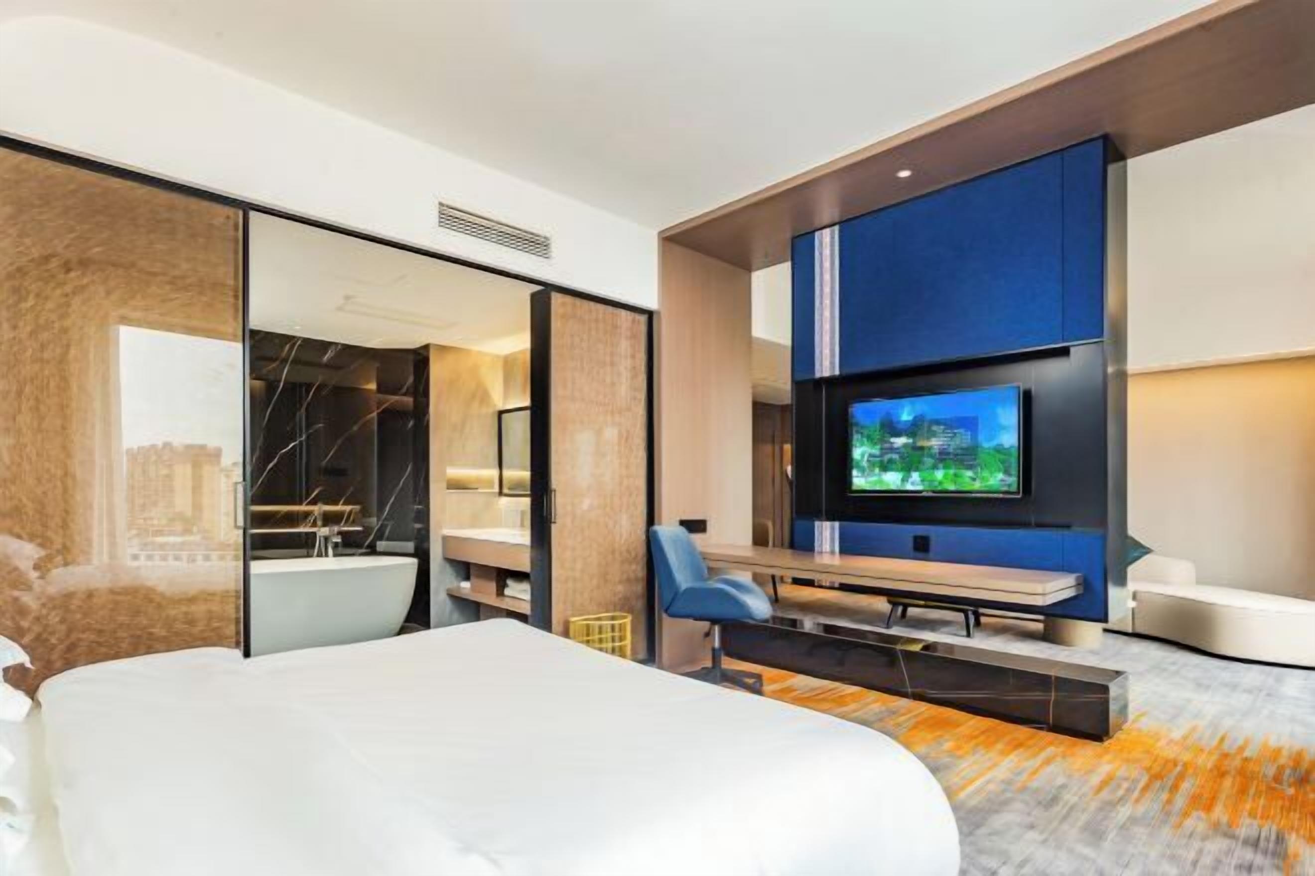 A modern hotel room with a large bed, a flat-screen TV mounted on the wall, a seating area with a bench, and a view of the cityscape through the glass door.