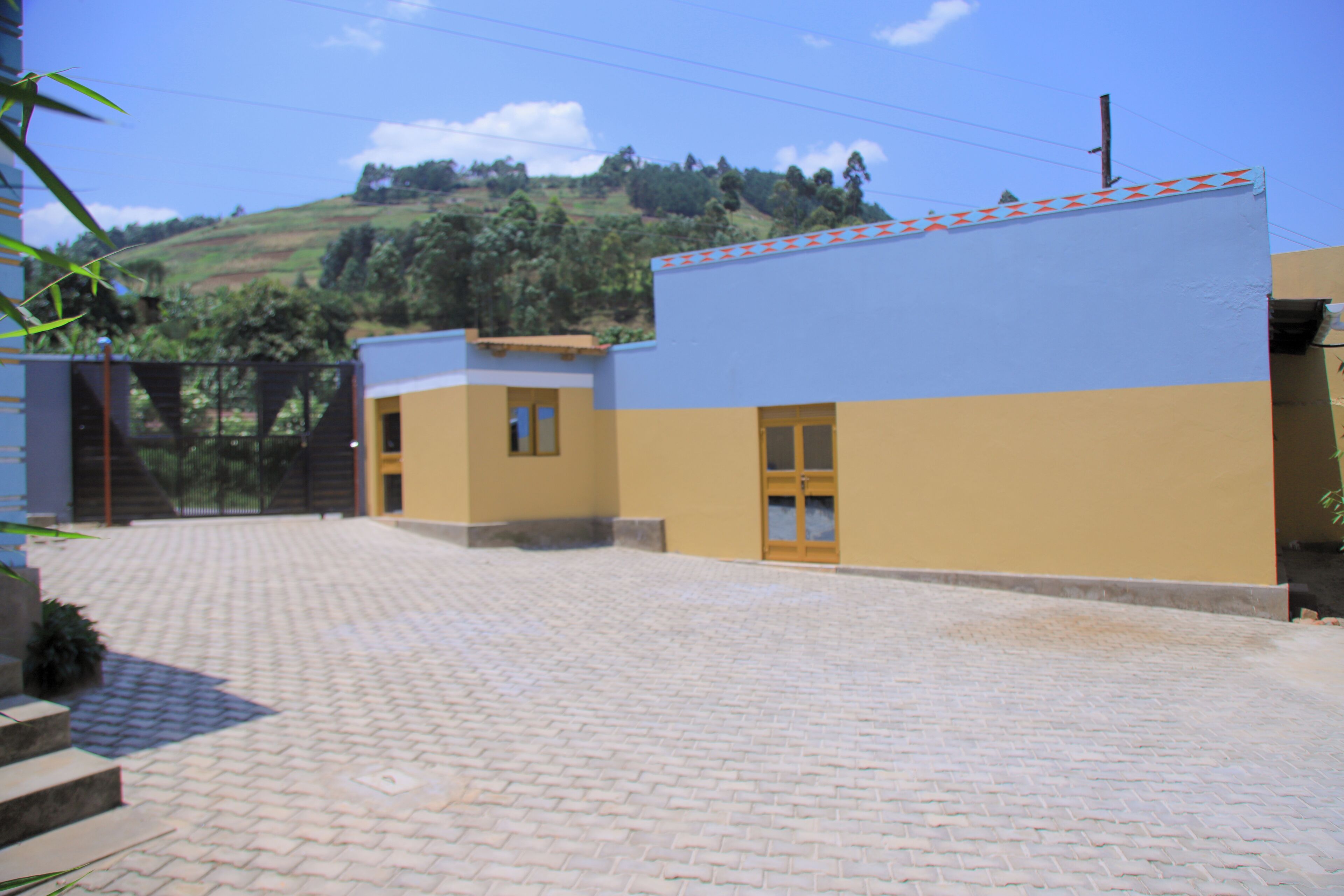 A paved courtyard with a building featuring a blue and yellow facade, a gate, and a hilly landscape in the background.