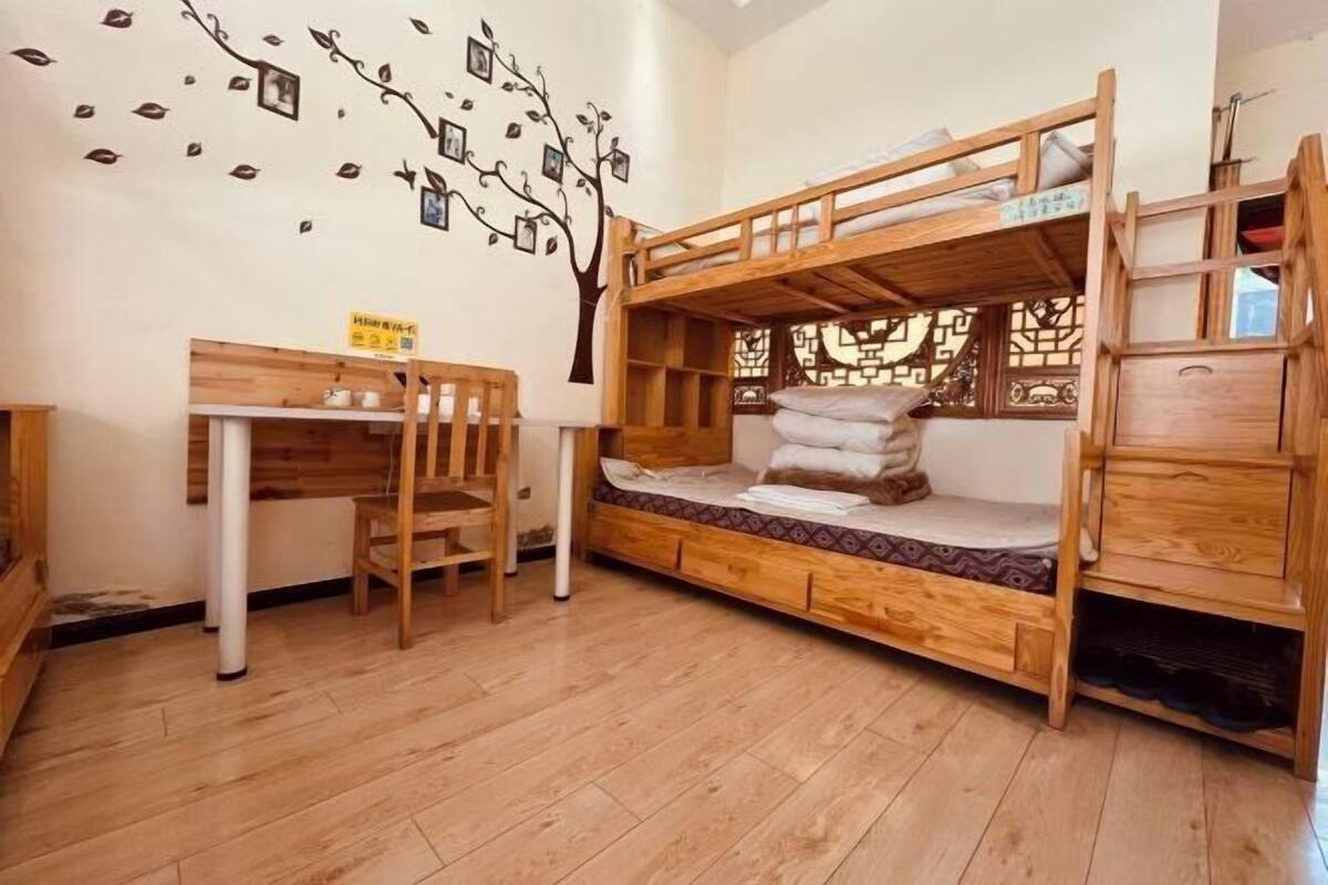 Ein Stockbett mit Schreibtisch und Stuhl, eine Wanddekoration mit Baummotiv und ein Fenster mit dekorativem Sichtschutz.