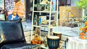 Un fauteuil en cuir noir, une bibliothèque blanche contenant divers objets et une table recouverte d’une nappe blanche, sur laquelle est posé un vase de fleurs.