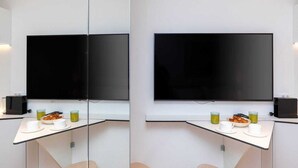 Una habitación moderna con un televisor de pantalla plana grande montado en la pared, un separador de vidrio y una mesa de desayuno con tazas y un plato de comida.