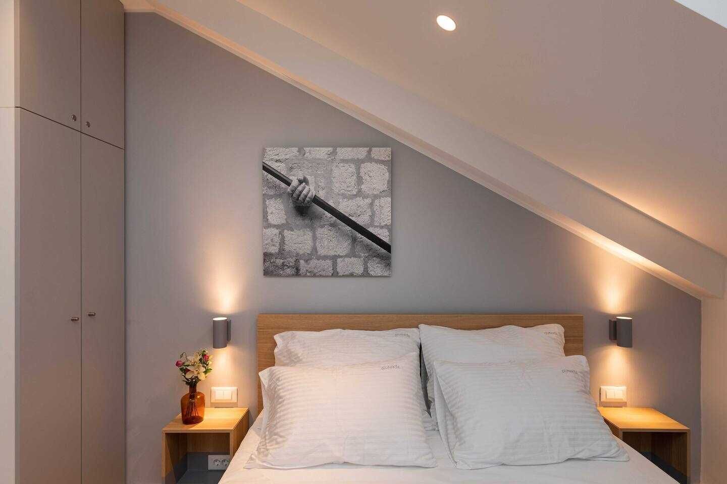 Een slaapkamer met een schuine plafond, een bed met wit beddengoed, twee nachtkastjes met lampen, en een wanddecoratie van een hand die een hamer vasthoudt.