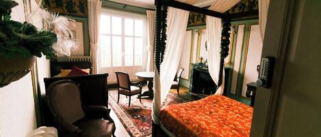 Una cama con dosel y cuatro columnas cubierta con una colcha floral, una silla de madera, una mesita pequeña y una chimenea en una habitación con paredes verdes y un ventanal grande.