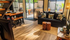 Uma sala de estar moderna com piso de madeira, um sofá preto com almofadas tropicais, uma mesa de centro de madeira e uma escada que leva a uma cozinha.