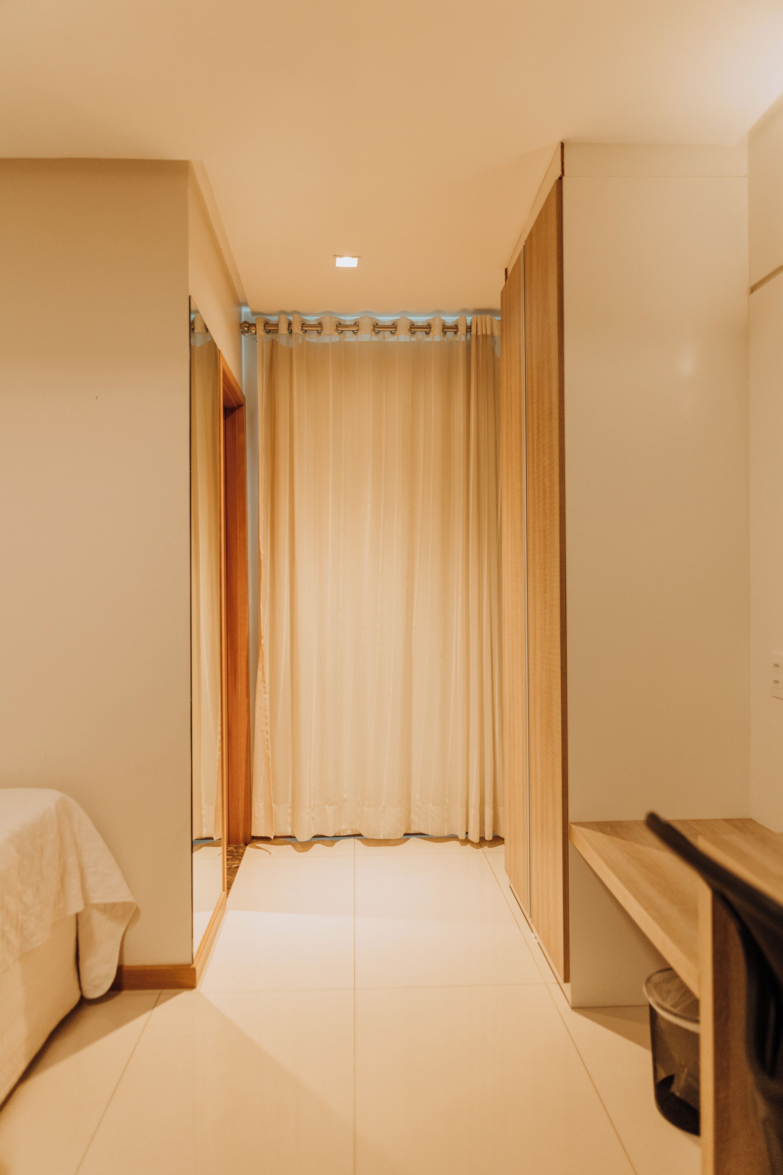 A hotel room with a bed, a wooden bench, a trash bin, and a curtain.