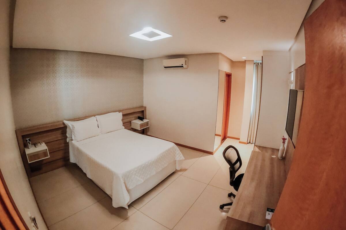 Quarto de hotel com cama, mesa de cabeceira, escrivaninha e uma secretária com cadeira.