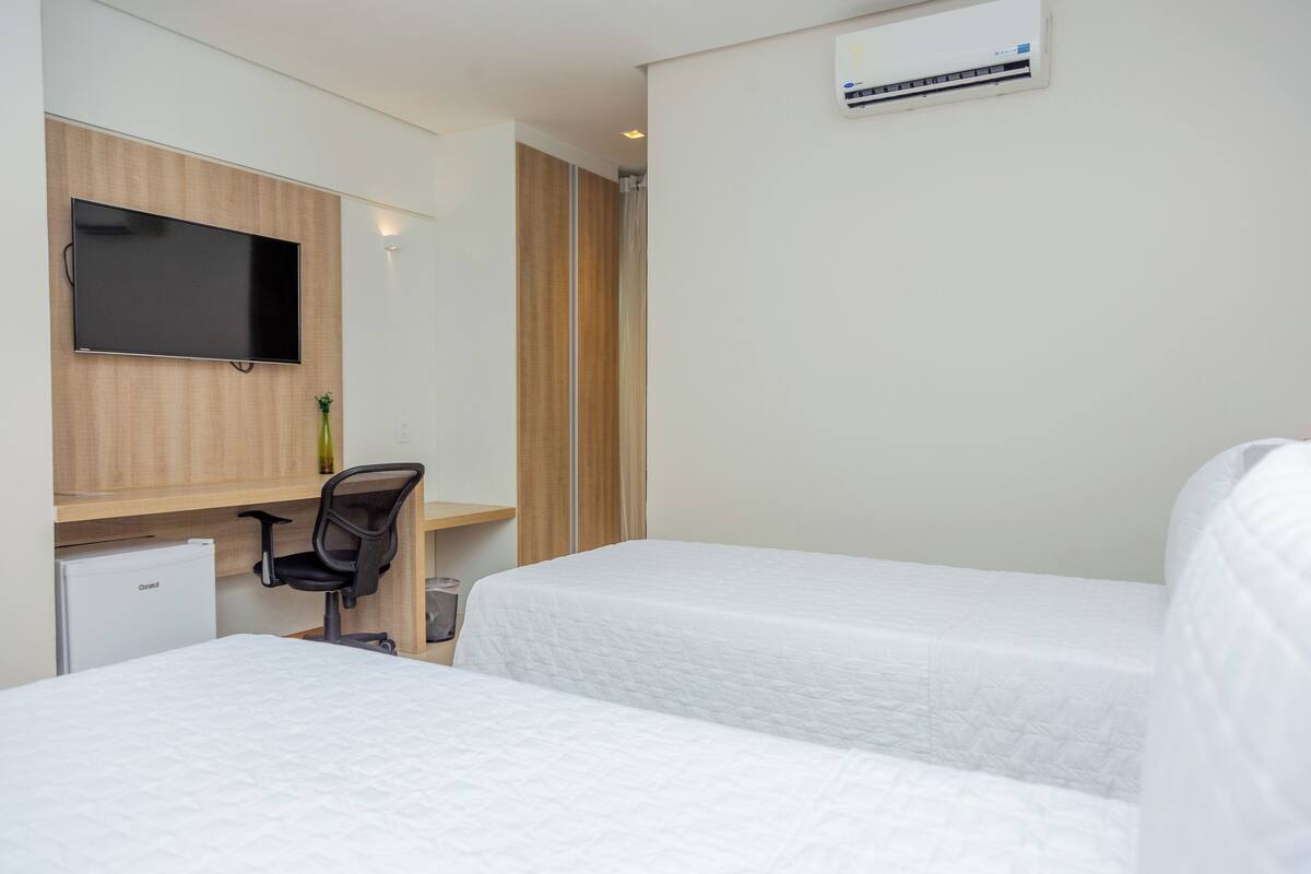 Quarto de hotel com duas camas, uma escrivaninha com cadeira, televisão e unidade de ar condicionado.