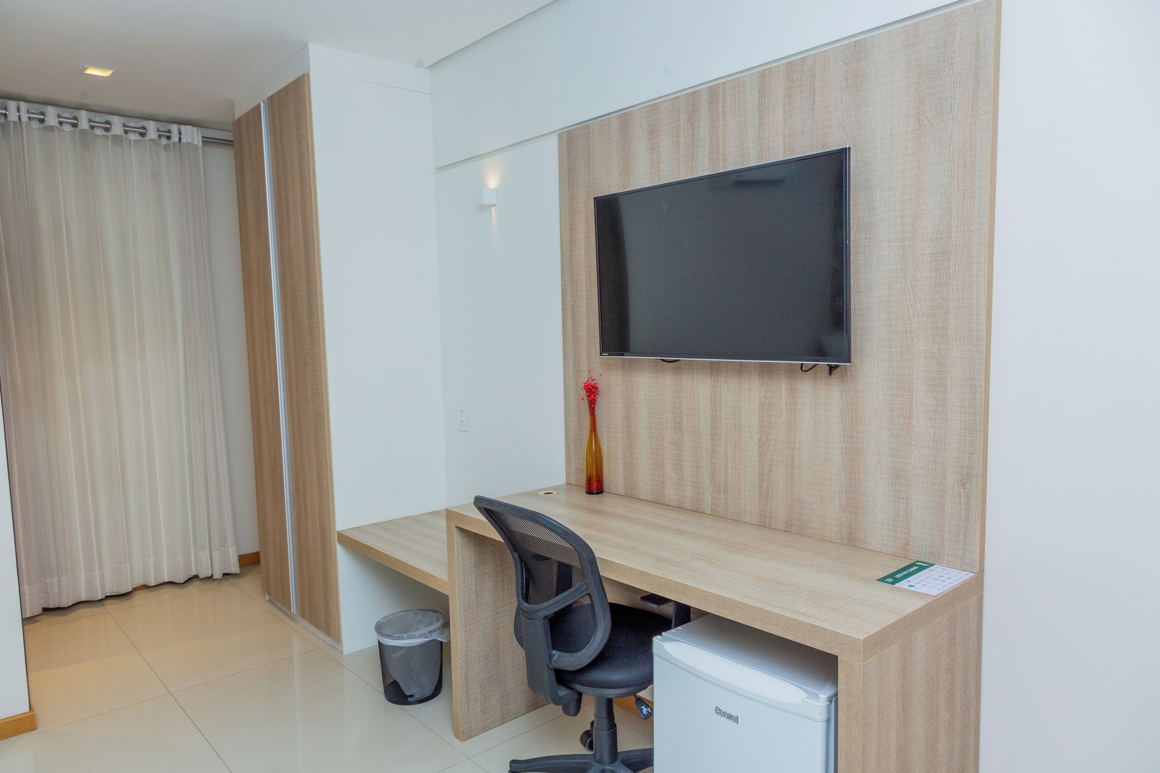 Ein moderner Arbeitsbereich mit einem Holztisch, einem schwarzen Bürostuhl, einem an der Wand befestigten Flachbildfernseher, einer kleinen Mülltonne und einem weißen Kühlschrank.