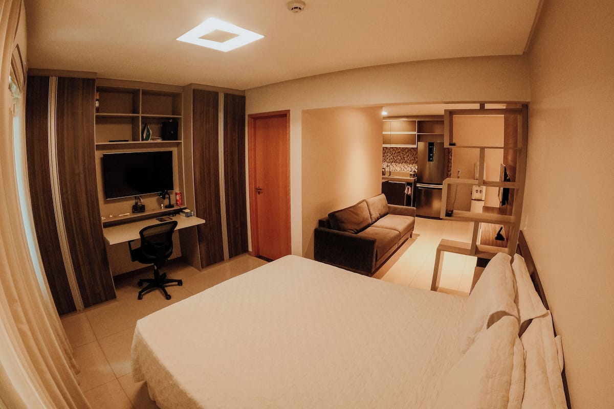 Quarto de hotel com cama, escrivaninha com computador, cadeira, sofá e cozinha compacta ao fundo.