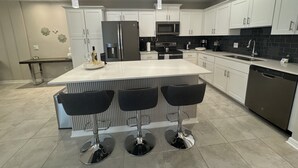 A modern kitchen with white cabinets, a central island with bar stools, stainless steel appliances, and pendant lighting. - 4731-103 Stay Magical • 2BR with Mickey Room & More (Kissimmee)