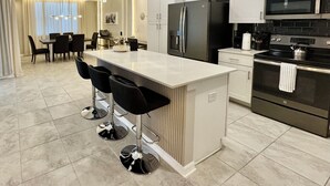 A modern kitchen with a central island, bar stools, stainless steel appliances, and a dining area with a table and chairs. - 4731-103 Stay Magical • 2BR with Mickey Room & More (Kissimmee)
