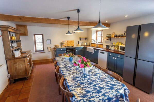 A kitchen with wooden cabinets, a refrigerator, and a dining table with a blue tablecloth and flowers. - Gorgeous spacious converted barn perfect for families with large garden. (Over Stowey)