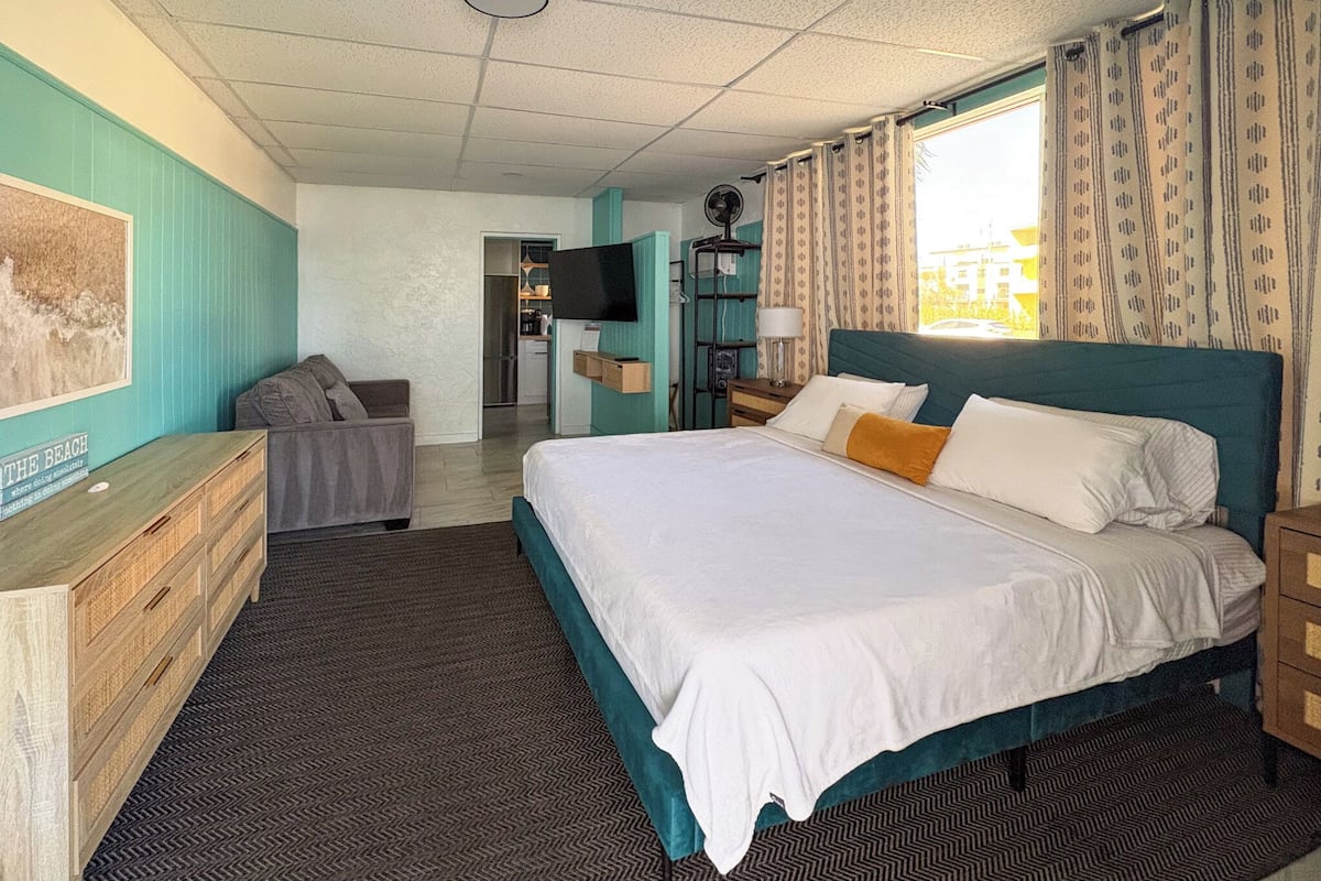 A hotel room with a bed, a desk, a chair, a TV, and a view of the outside.