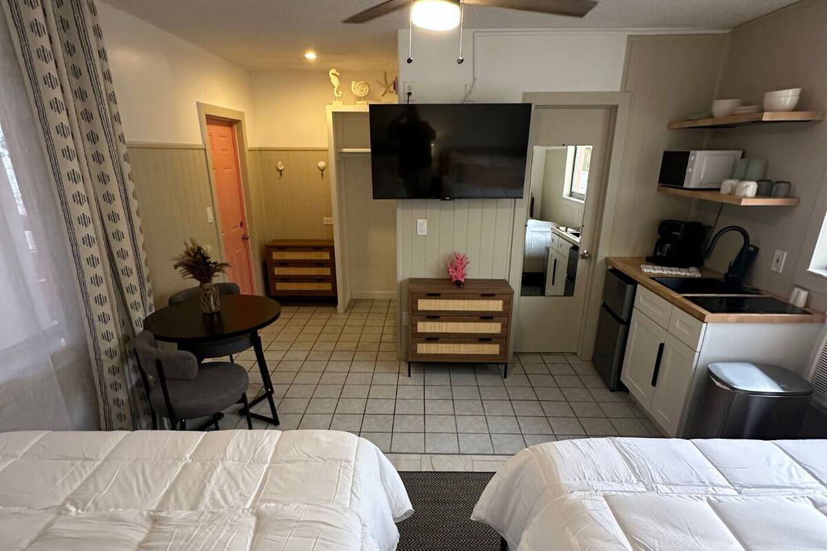 A hotel room with two beds, a small dining table, a kitchenette, a TV, and a bathroom visible through an open door.