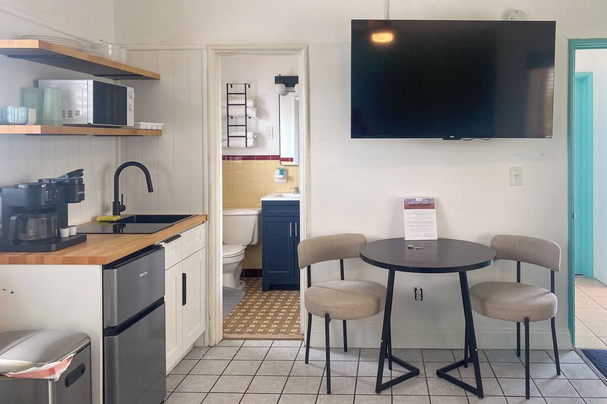 A compact kitchen with a microwave, coffee maker, and a refrigerator. There is a dining area with a table and four chairs. A flat-screen TV is mounted on the wall above the dining table.