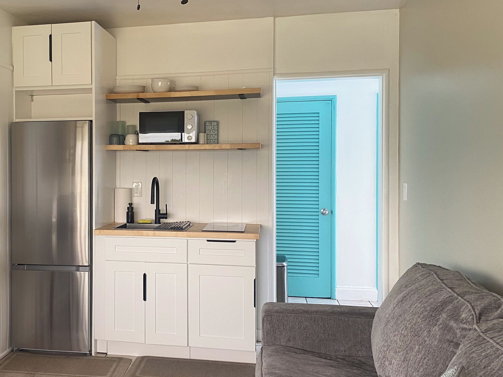 Una cocina compacta con armarios blancos, un refrigerador de acero inoxidable, un microondas y un lavamanos. Hay una puerta color turquesa que da a otra habitación.