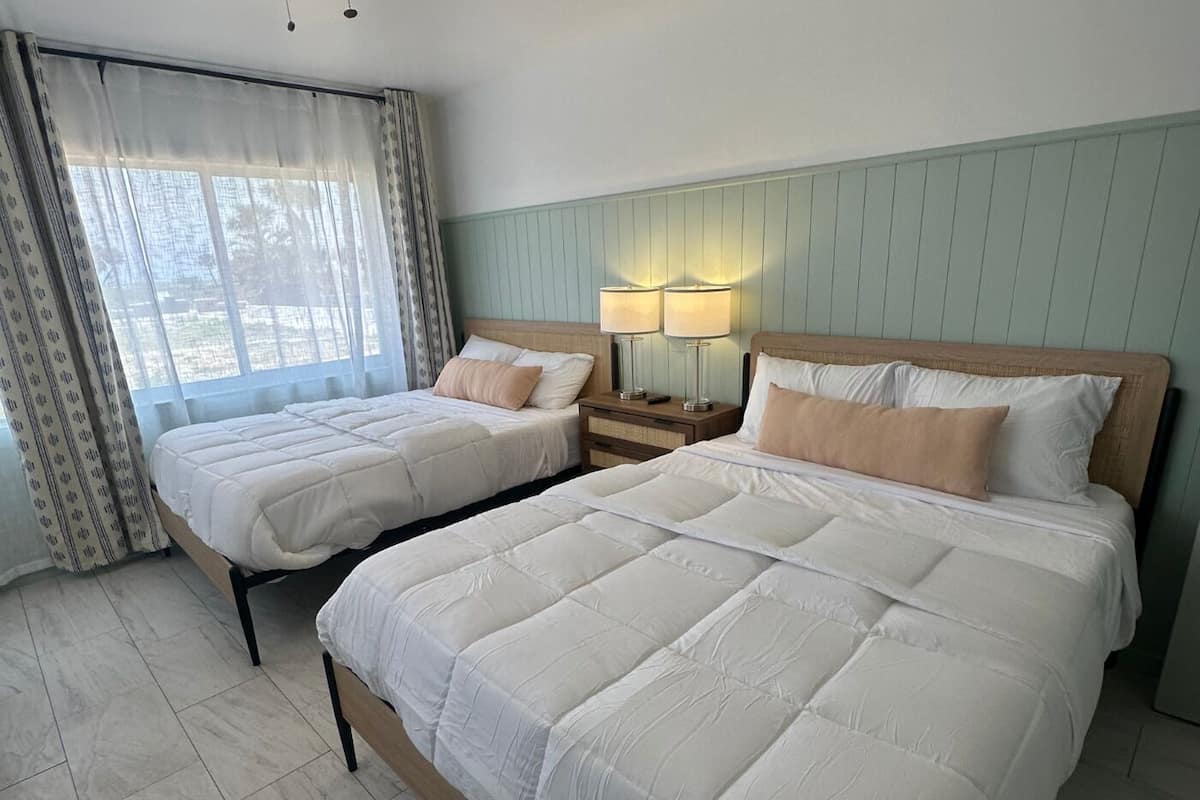 A hotel room with two beds, a nightstand with lamps, and a window with curtains.