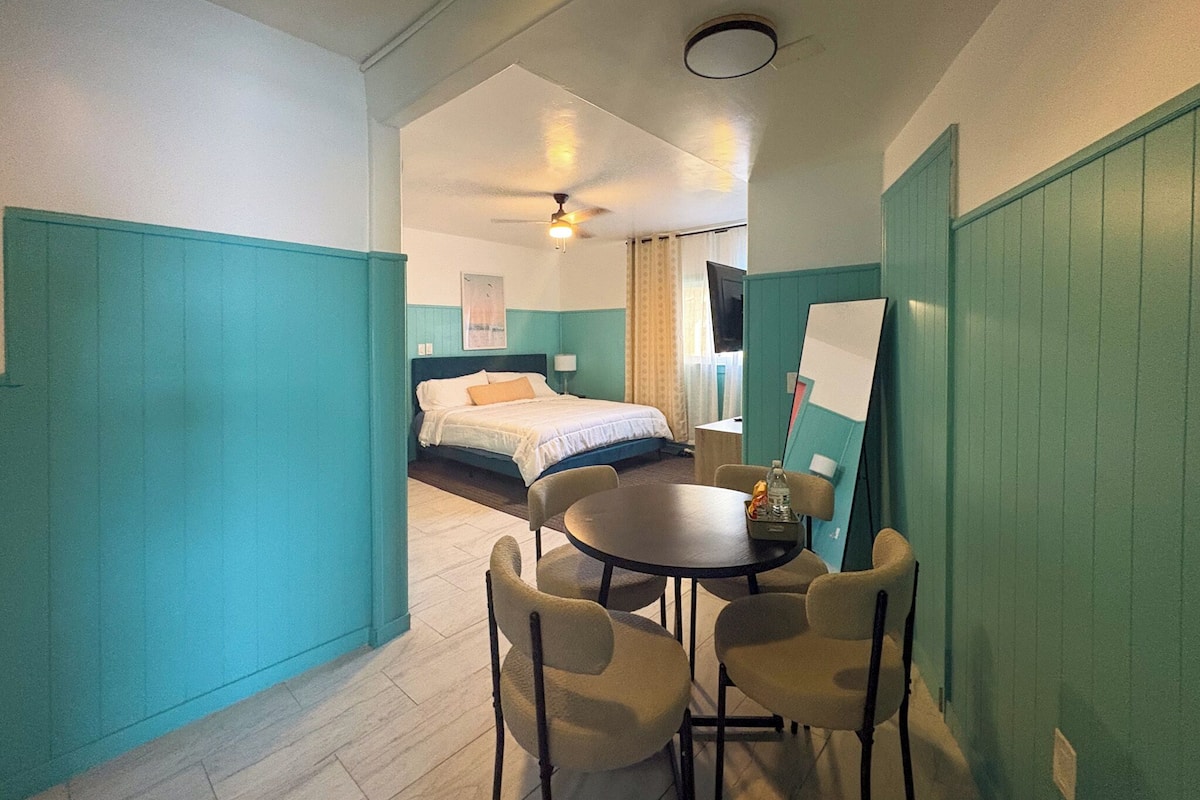 A hotel room with a bed, a desk with chairs, a ceiling fan, and a view of another room.
