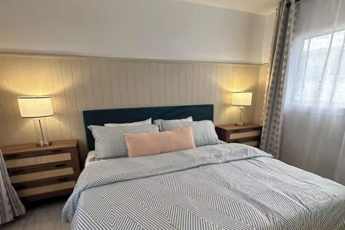 A bedroom with a bed, two bedside lamps, a ceiling fan, and a window with curtains.
