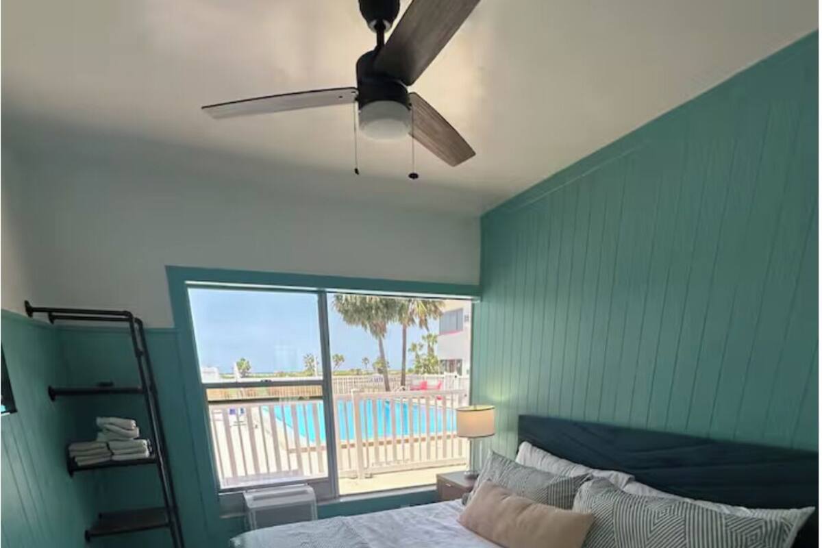 A bedroom with a bed, a ceiling fan, a window with a view of a pool, and a balcony.