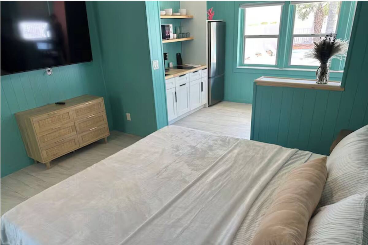 A bedroom with a bed, a dresser, a TV mounted on the wall, a window with a view, and a small shelf with items.