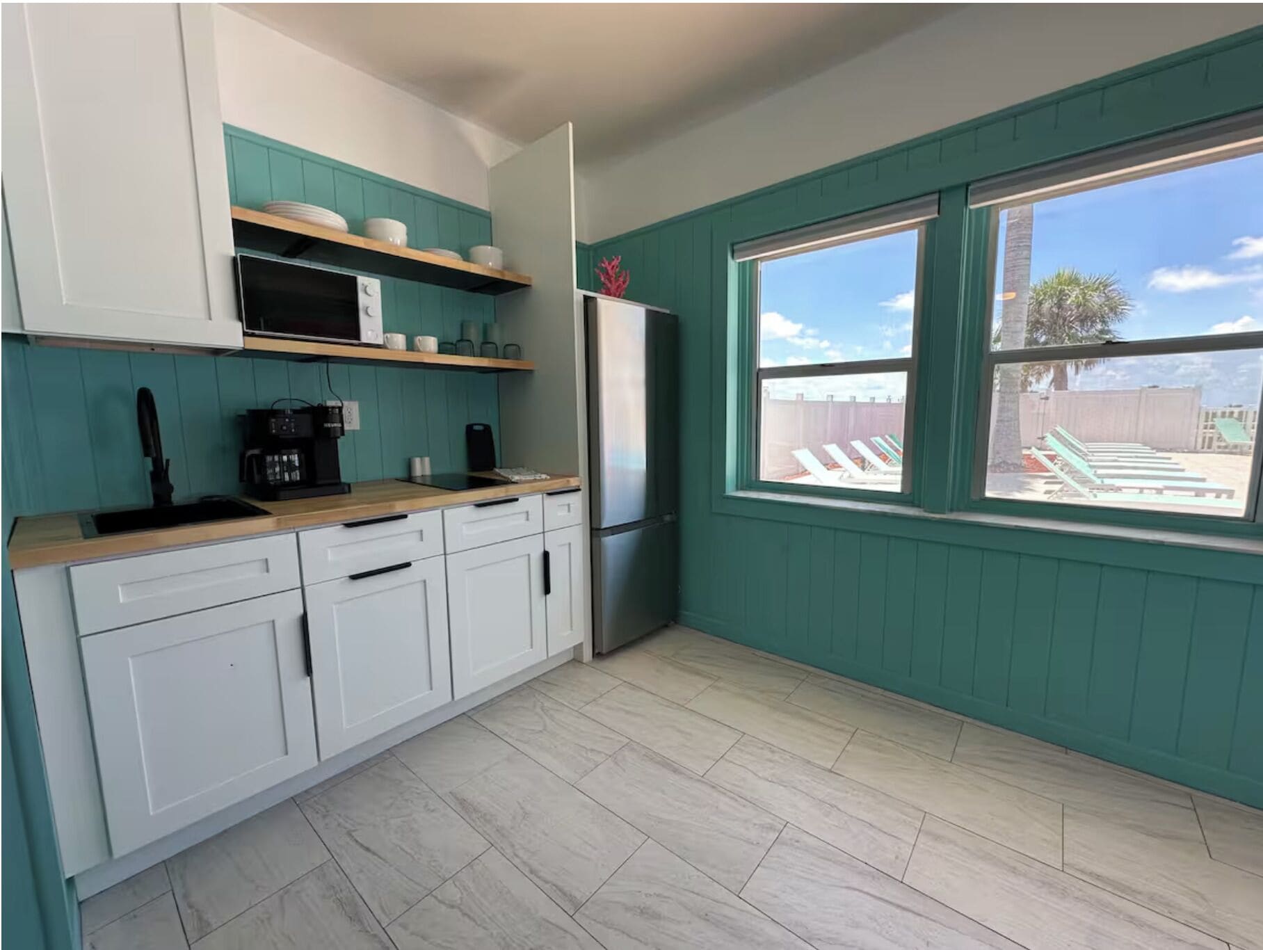 A kitchen with white cabinets, a wooden countertop, a microwave, a refrigerator, and a window with a view of a backyard.