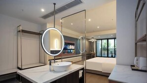 Un baño de hotel moderno con un espejo grande, un espejo redondo, un lavabo blanco y una cama.