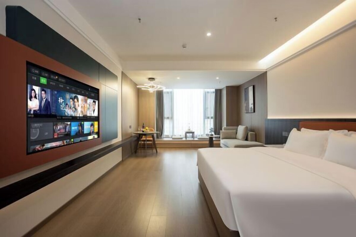 A hotel room with a large flat-screen TV mounted on the wall displaying various channels, a bed with white linens, a small table with a lamp, and a view of the room's interior through a window.