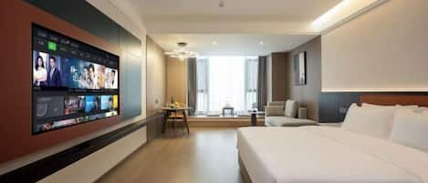 Habitación de hotel con un televisor de pantalla plana grande montado en la pared mostrando varios canales, una cama con ropa blanca, una mesita de noche con lámpara, y vista al interior de la habitación desde una ventana.